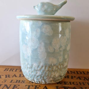 May include: A ceramic jar with a lid and a bird-shaped handle. The jar is light blue with white and brown accents. The jar is cylindrical and has a textured bottom. The lid is round and sits on top of the jar.