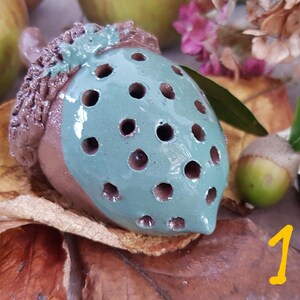 May include: A ceramic acorn with a brown cap and a teal body with holes. The acorn is sitting on a bed of brown leaves.