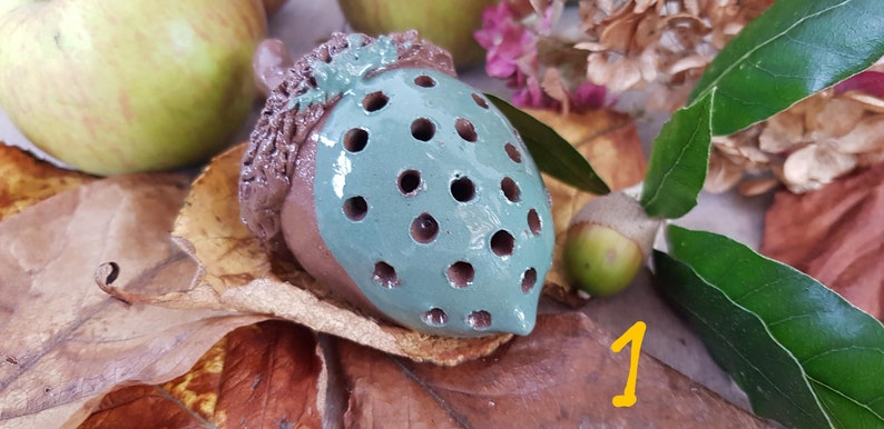 May include: A ceramic acorn with a brown cap and a teal body with holes. The acorn is sitting on a bed of brown leaves.