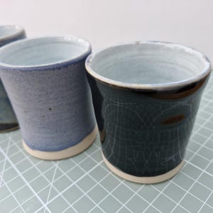 May include: Handmade ceramic cups in blue and green hues. The cups feature a distinctive crackle glaze, with a light-coloured rim and base. They are presented on a light green grid surface.