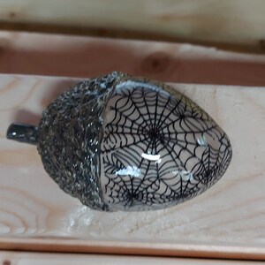 May include: A ceramic acorn with a black and white spiderweb design. The acorn is glazed and has a textured surface.
