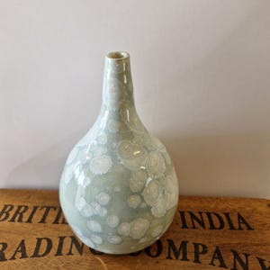 May include: A pale green ceramic vase with a rounded body and a narrow neck. The vase features a speckled, crystalline pattern in white, giving it a textured look. The vase is placed on a wooden surface.