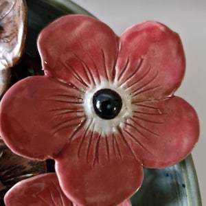May include: Close-up of a ceramic floral design. The flowers are a muted red with a black centre and white accents. The petals have detailed lines. The background is a muted green and blue.