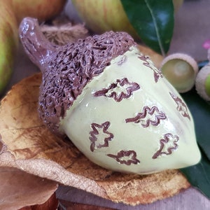 May include: A ceramic acorn ornament with a brown cap and a light green body decorated with brown leaf designs. The acorn is sitting on a brown leaf and a green leaf. The number 4 is written in yellow on a green leaf.