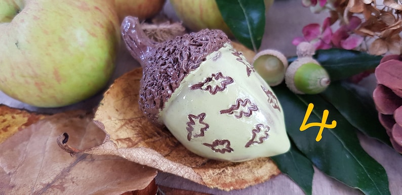 May include: A ceramic acorn ornament with a brown cap and a light green body decorated with brown leaf designs. The acorn is sitting on a brown leaf and a green leaf. The number 4 is written in yellow on a green leaf.