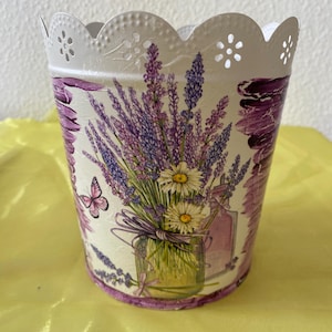 Könnte beinhalten: Weißer Metalltopf mit einem gewellten Rand und einem Lavendel-Blumendesign. Der Topf zeigt einen Strauß Lavendelblüten, Gänseblümchen und ein Glasgefäß mit einem rosa Band.