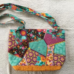 May include: A patchwork tote bag with a floral and geometric pattern. The bag features a variety of fabrics in teal, orange, pink, and black. The bag has a matching strap and a yellow-orange bottom.