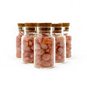 Puede incluir: Seis pequeños frascos de vidrio llenos de piedras preciosas rosas, cada uno sellado con un tapón de corcho. Los frascos están dispuestos en semicírculo sobre un fondo blanco. Las piedras tienen una forma suave y redondeada.