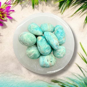 Pu&ograve; includere: Una ciotola bianca piena di pietre di agata rotolata di colore blu turchese e bianco. Le pietre sono lisce e levigate e hanno un aspetto naturale e organico.