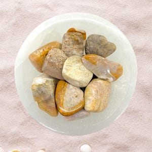 Peut inclure: Un ensemble de pierres polies aux tons de beige, marron et orange, nich&eacute;es dans un bol blanc peu profond. Les pierres varient en taille et en forme, avec des surfaces lisses et arrondies. Le bol repose sur une surface rose clair.