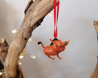 May include: A red and orange plastic ornament of Pumbaa the warthog from Disney's The Lion King, hanging from a red ribbon on a branch.