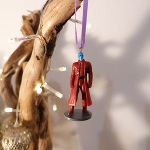 May include: A blue-skinned figure in a red robe with a purple ribbon hanging from the top of its head. The figure is a Christmas ornament.
