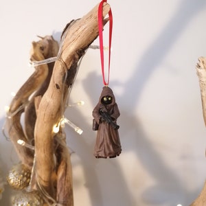 May include: A brown and black Star Wars character ornament hanging from a red ribbon on a branch with white lights. The character is wearing a brown robe and holding a weapon.