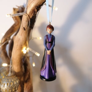 May include: Purple and black Christmas ornament featuring a woman with brown hair wearing a purple dress and black cape.