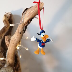 May include: A yellow and blue plastic Donald Duck Christmas ornament with a red bow hanging from a driftwood branch with white lights.