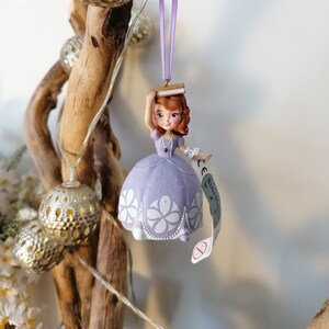 May include: Purple and white princess ornament with a book on her head. The ornament is hanging from a purple ribbon. The ornament has a tag that says "Disney Princess".