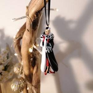Puede incluir: Un adorno navideño con Cruella de Vil de la película de Disney 101 Dálmatas. El adorno está hecho de plástico y está pintado en negro, blanco y rojo. Cruella lleva un abrigo negro con un cuello de piel blanca y un vestido rojo. Lleva un manguito de piel blanca en su mano derecha.