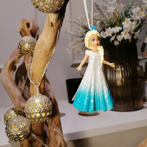 Peut inclure: Une poup&eacute;e en plastique blanche et bleue avec une tresse blonde suspendue &agrave; une ficelle blanche. La poup&eacute;e porte une robe blanche avec une jupe bleu d&eacute;grad&eacute; et une couronne bleue. La poup&eacute;e est suspendue &agrave; une branche avec des guirlandes lumineuses.