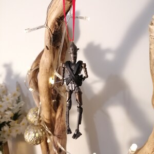 Puede incluir: Un adorno de robot met&aacute;lico gris y marr&oacute;n con una cinta roja colgando de una rama. El robot tiene una X negra en el pecho y lleva un casco dorado.