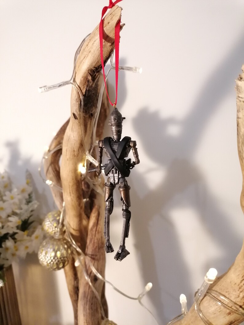 Puede incluir: Un adorno de robot met&aacute;lico gris y marr&oacute;n con una cinta roja colgando de una rama. El robot tiene una X negra en el pecho y lleva un casco dorado.