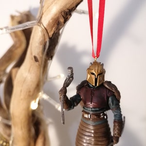 May include: A gold and brown Mandalorian ornament with a red ribbon hanging from a branch. The ornament is holding a silver weapon.