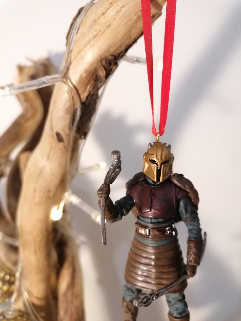 May include: A gold and brown Mandalorian ornament with a red ribbon hanging from a branch. The ornament is holding a silver weapon.