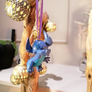 Pode incluir: Um ornamento de Stitch azul e turquesa pendurado em uma fita roxa em um galho de madeira com l&acirc;mpadas douradas.