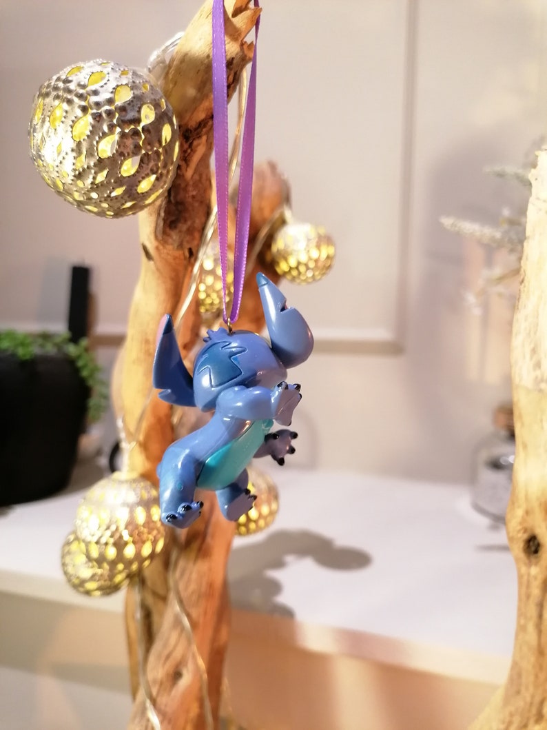 Pode incluir: Um ornamento de Stitch azul e turquesa pendurado em uma fita roxa em um galho de madeira com l&acirc;mpadas douradas.