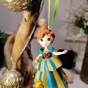 May include: A green ribbon hangs from a branch, holding a small plastic figurine of a girl with red hair wearing a blue and gold dress. The girl is holding a small yellow bird in her hand.