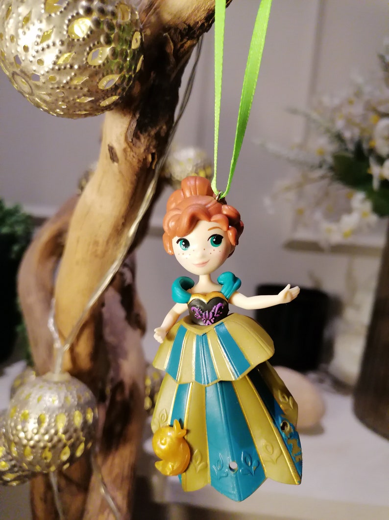 May include: A green ribbon hangs from a branch, holding a small plastic figurine of a girl with red hair wearing a blue and gold dress. The girl is holding a small yellow bird in her hand.