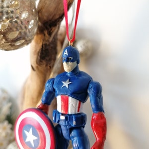 May include: A blue and red Captain America action figure ornament with a red ribbon and a shield. The figure is wearing a blue and red costume with a white star on the chest and a white star on the shield.