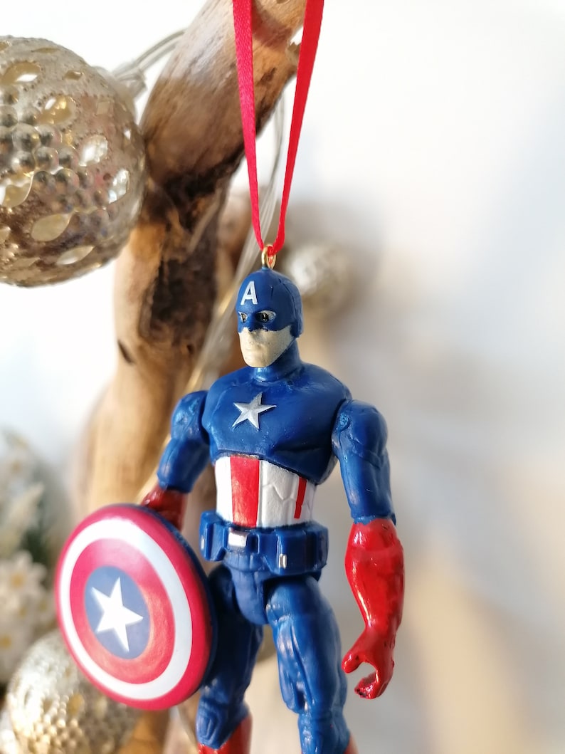 May include: A blue and red Captain America action figure ornament with a red ribbon and a shield. The figure is wearing a blue and red costume with a white star on the chest and a white star on the shield.