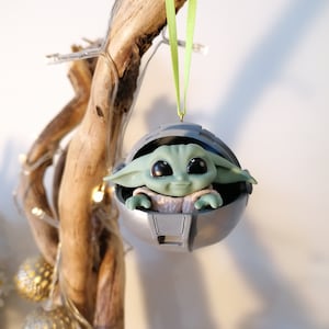 May include: A green and gray Christmas ornament featuring a character from the Star Wars franchise, The Mandalorian, known as "The Child" or "Baby Yoda", sitting inside a pod. The ornament is hanging from a green ribbon.