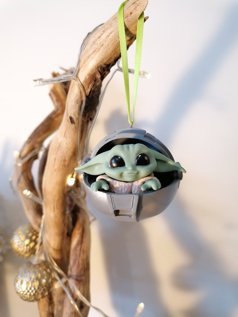 May include: A green and gray Christmas ornament featuring a character from the Star Wars franchise, The Mandalorian, known as "The Child" or "Baby Yoda", sitting inside a pod. The ornament is hanging from a green ribbon.