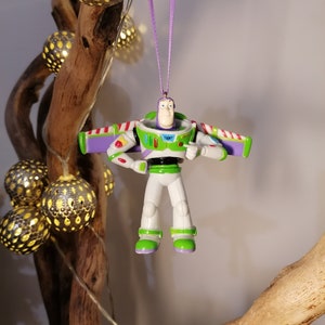 Peut inclure: Ornement de No&euml;l Buzz l'&Eacute;clair, avec un design de combinaison spatiale blanche, verte et violette. Suspendu par un ruban violet. Le fond comprend des lumi&egrave;res d&eacute;coratives et une branche en bois.