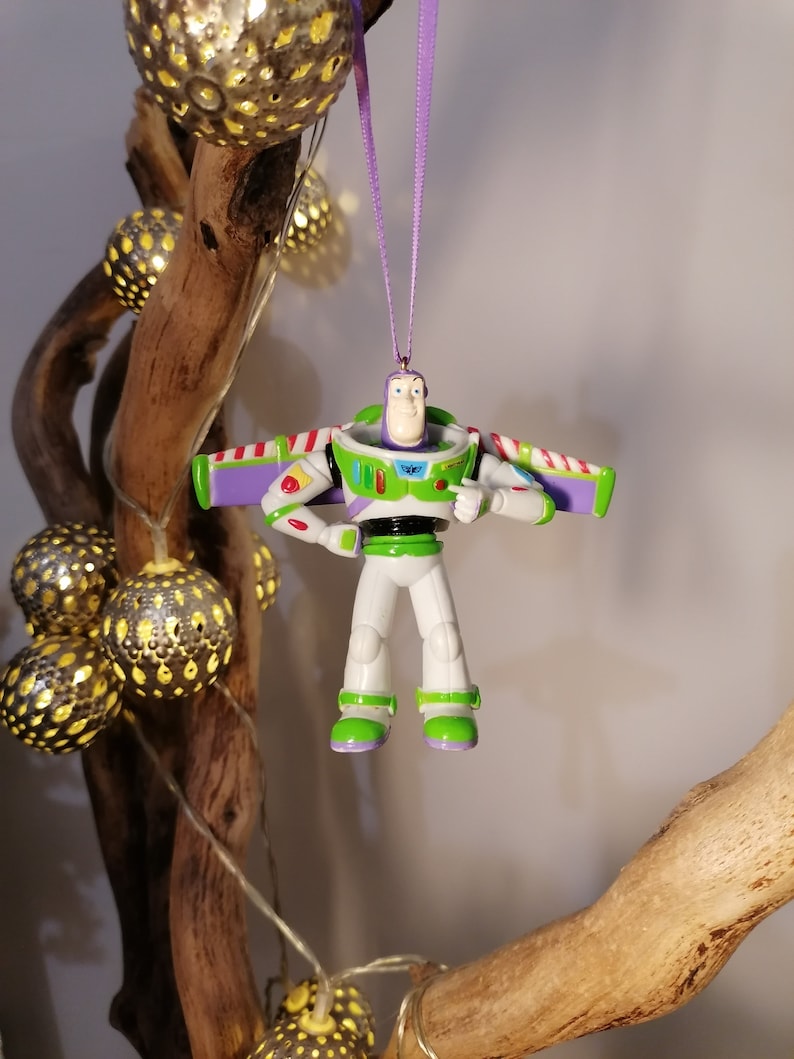 Peut inclure: Ornement de No&euml;l Buzz l'&Eacute;clair, avec un design de combinaison spatiale blanche, verte et violette. Suspendu par un ruban violet. Le fond comprend des lumi&egrave;res d&eacute;coratives et une branche en bois.