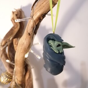 May include: A green and blue Christmas ornament featuring a character from the Star Wars franchise. The ornament is hanging from a branch with a green ribbon.