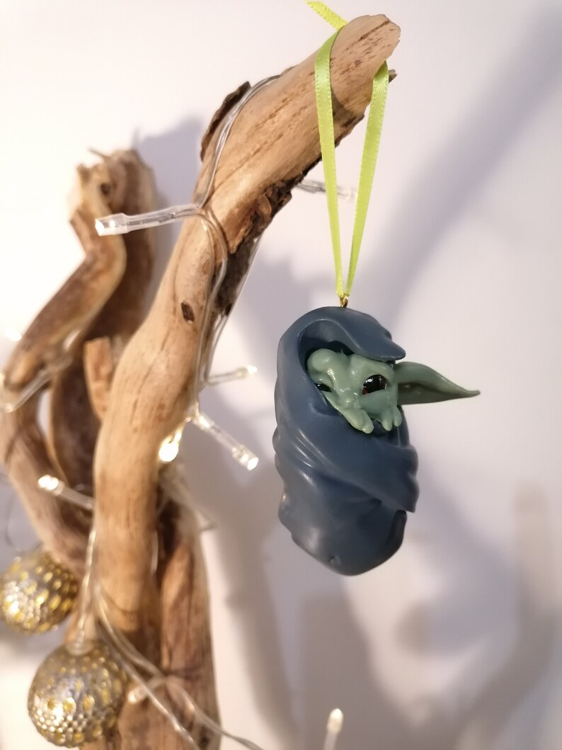 May include: A green and blue Christmas ornament featuring a character from the Star Wars franchise. The ornament is hanging from a branch with a green ribbon.