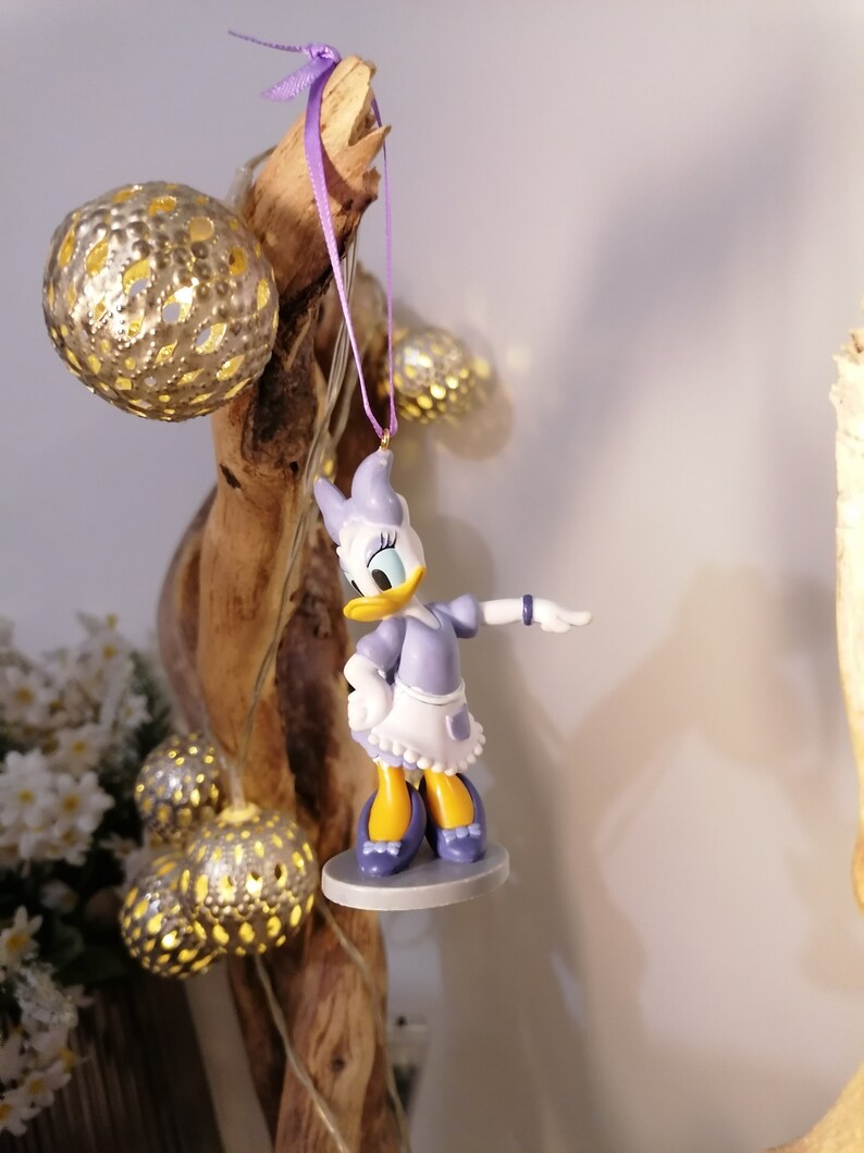 Puede incluir: Figurita de pato de dibujos animados de color morado y blanco que lleva un vestido y sostiene una flor morada. La figurita est&aacute; colgada de una rama con una cinta morada.