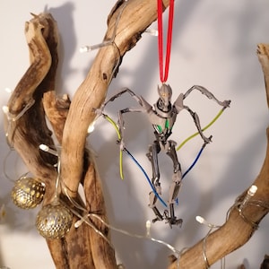 May include: A gray and green robot Christmas ornament hanging from a branch with a red ribbon. The robot has long arms and legs and is posed in a dynamic action pose. The ornament is surrounded by white lights.