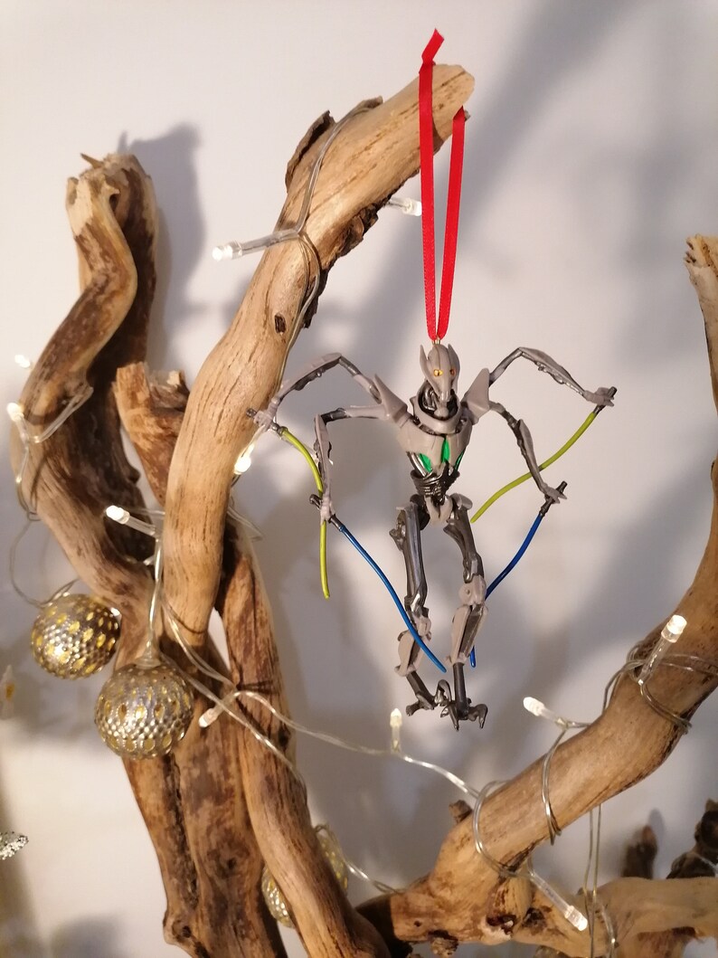 May include: A gray and green robot Christmas ornament hanging from a branch with a red ribbon. The robot has long arms and legs and is posed in a dynamic action pose. The ornament is surrounded by white lights.