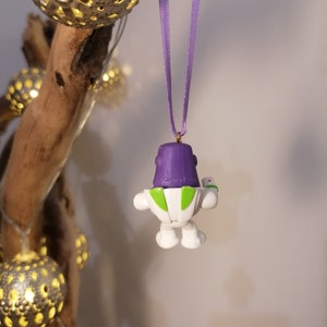 May include: Purple and white plastic ornament of Buzz Lightyear from the Toy Story franchise hanging from a purple ribbon. The ornament is attached to a gold hook.