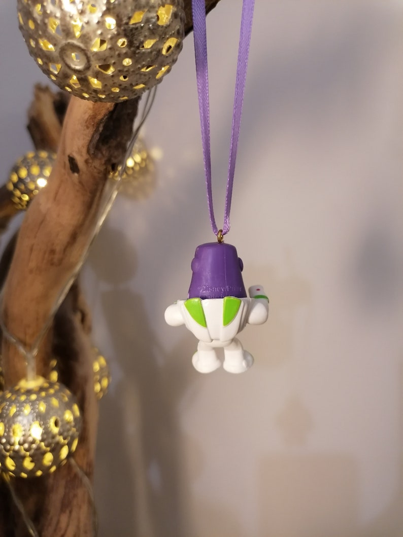 May include: Purple and white plastic ornament of Buzz Lightyear from the Toy Story franchise hanging from a purple ribbon. The ornament is attached to a gold hook.