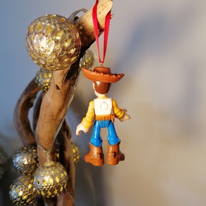 May include: A plastic toy ornament of Woody from Toy Story, wearing his signature brown hat, yellow shirt, blue jeans, and brown boots. The ornament is hanging from a red ribbon on a branch.