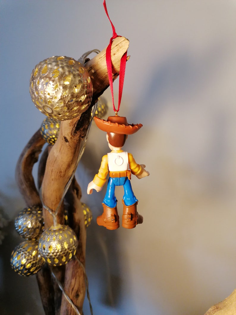 May include: A plastic toy ornament of Woody from Toy Story, wearing his signature brown hat, yellow shirt, blue jeans, and brown boots. The ornament is hanging from a red ribbon on a branch.