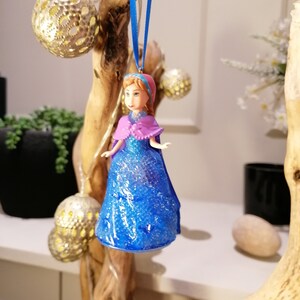 Puede incluir: Un adorno de vestido azul y morado con purpurina con una cabeza de mu&ntilde;eca que lleva un sombrero rosa. El adorno est&aacute; colgado de una cinta azul en una rama de madera.