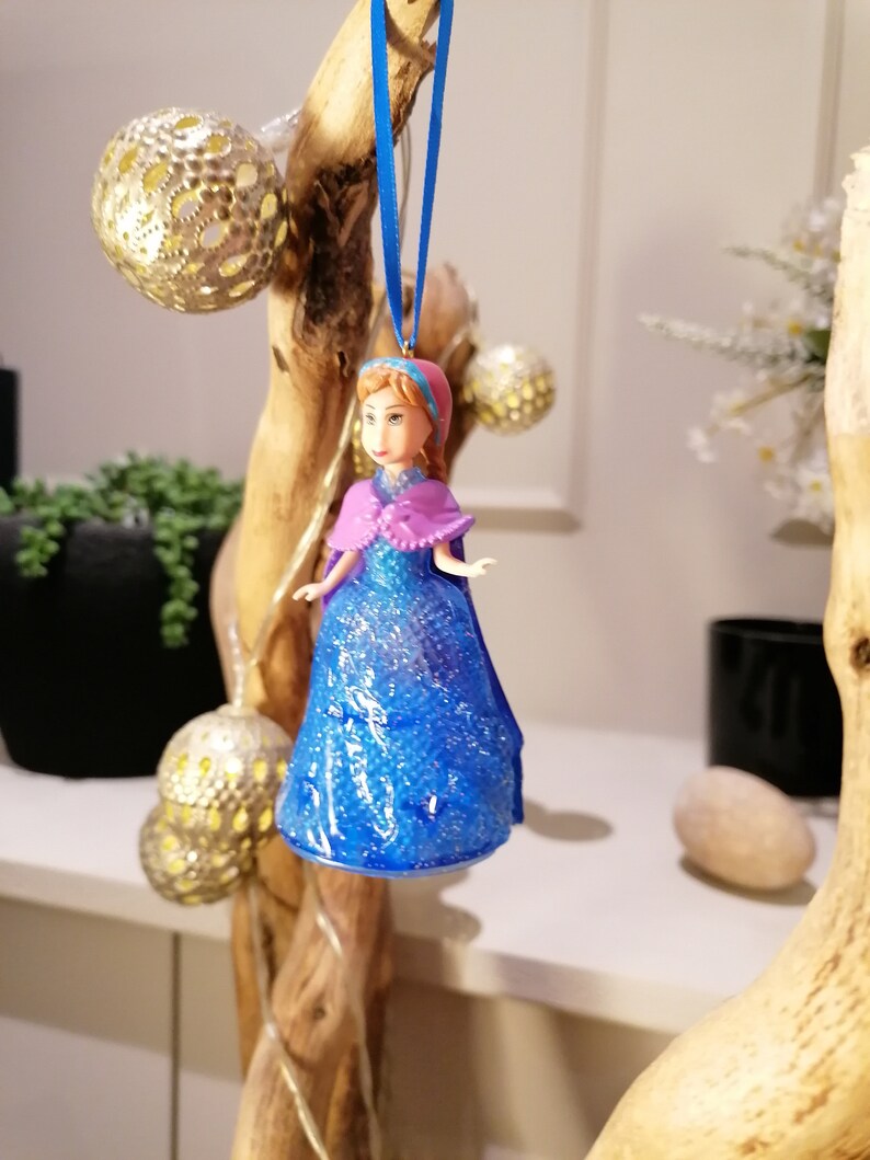 Puede incluir: Un adorno de vestido azul y morado con purpurina con una cabeza de mu&ntilde;eca que lleva un sombrero rosa. El adorno est&aacute; colgado de una cinta azul en una rama de madera.