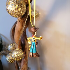 May include: A Woody action figure Christmas ornament, hanging from a yellow ribbon. The figure is wearing a brown cowboy hat, blue pants, and brown boots. Decorative gold-colored, spherical lights and a wooden branch are in the background.
