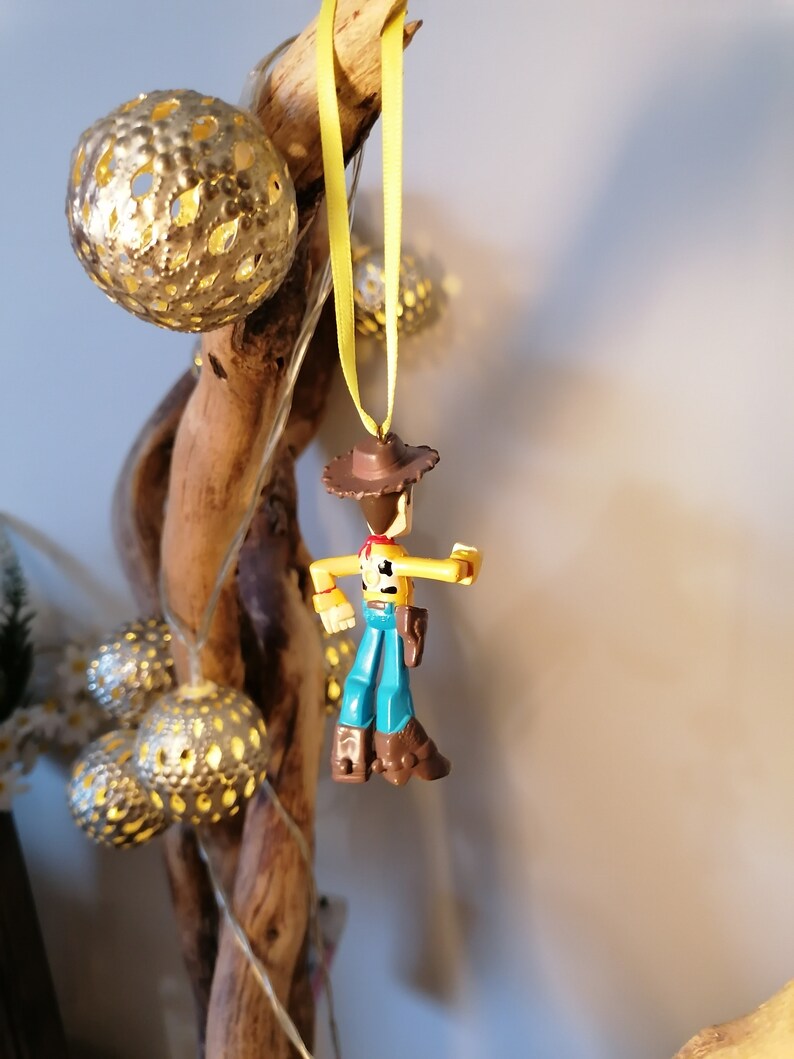 May include: A Woody action figure Christmas ornament, hanging from a yellow ribbon. The figure is wearing a brown cowboy hat, blue pants, and brown boots. Decorative gold-colored, spherical lights and a wooden branch are in the background.