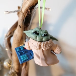 May include: A green and brown character from the Star Wars franchise, known as "The Child" or "Baby Yoda", is wearing a tan coat with a fur collar and holding a blue and silver device. The character is hanging from a branch with a green ribbon.